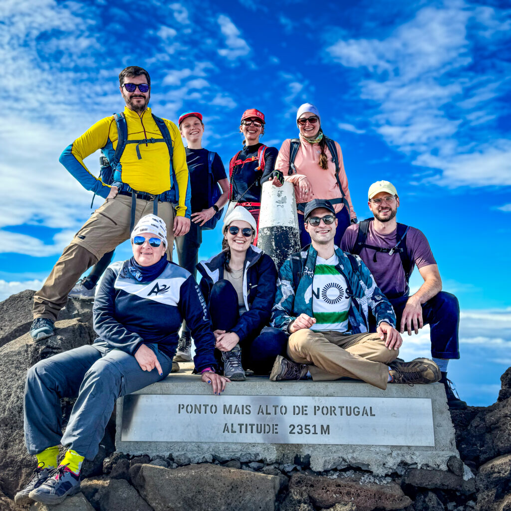 A group of hikers celebrating at the summit of Mount Pico, the highest point in Portugal, with a blue sky and panoramic views in the background.