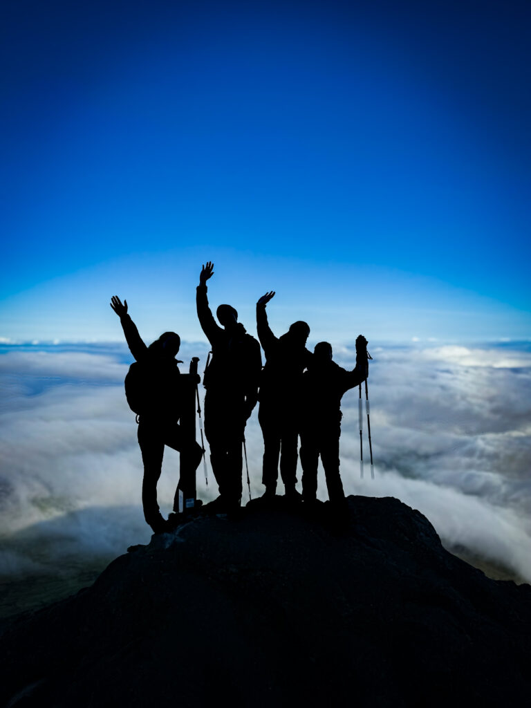 A group of hikers celebrates at the summit of Mount Pico, standing above the clouds with trekking poles raised in triumph.