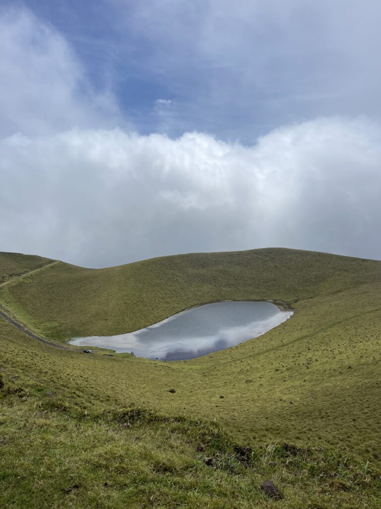 A secluded lagoon nestled within rolling green hills on Pico Island, with clouds floating above, creating a mystical atmosphere.
