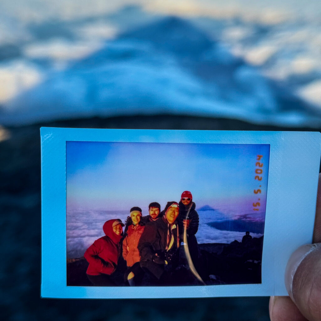 A hand holding a Polaroid photo of a group of hikers at the summit of Mount Pico, taken during sunrise after an overnight climb, with clouds and the mountain peak in the background.