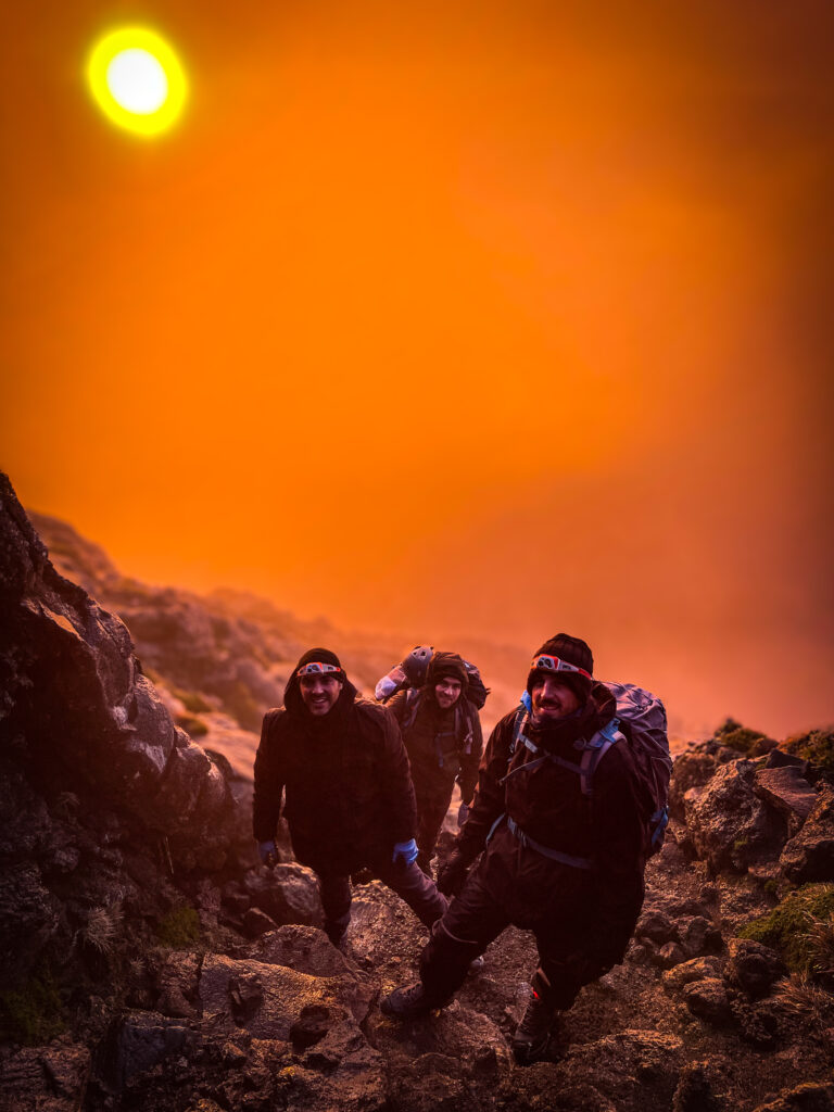 Three hikers ascending Mount Pico in the early morning, illuminated by a golden sunrise breaking through the mist.