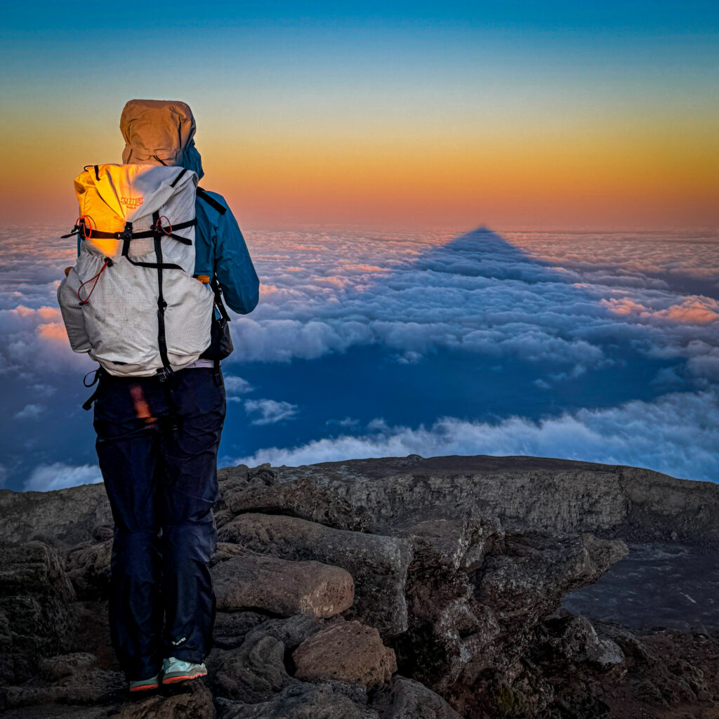 A hiker standing at the summit of Mount Pico at sunrise, gazing at the mountain's perfectly triangular shadow stretching across a sea of clouds.