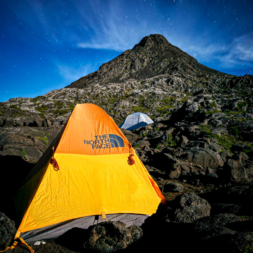 A brightly colored tent set up on the rocky slopes of Mount Pico under a starry night sky, with the mountain peak towering in the background.