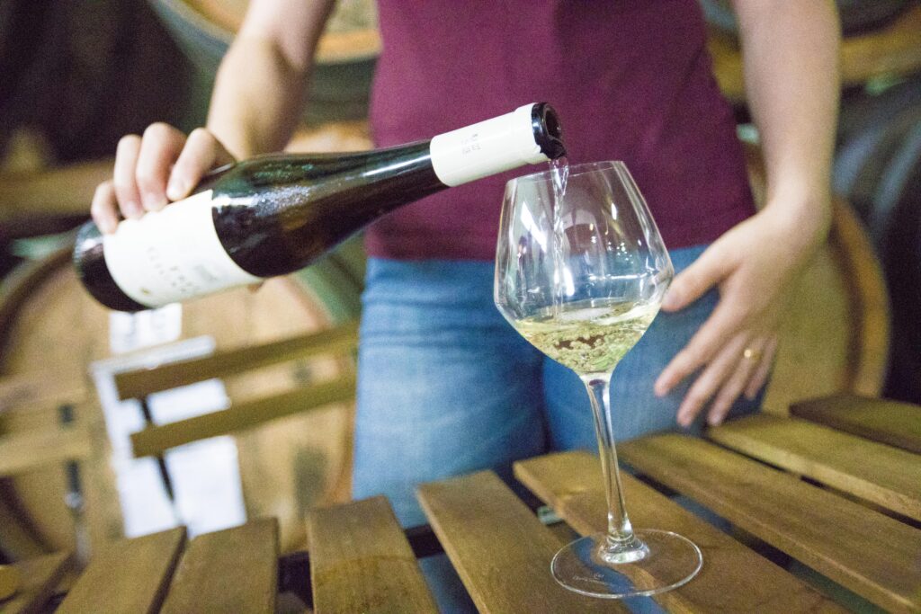 A person pouring a glass of white wine in a traditional wine cellar with wooden barrels in the background.