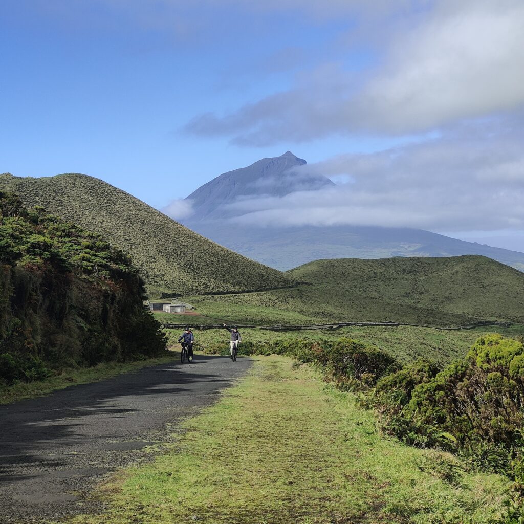 Two cyclists ride along a scenic volcanic trail with the majestic Pico Mountain in the background, partially covered by clouds.