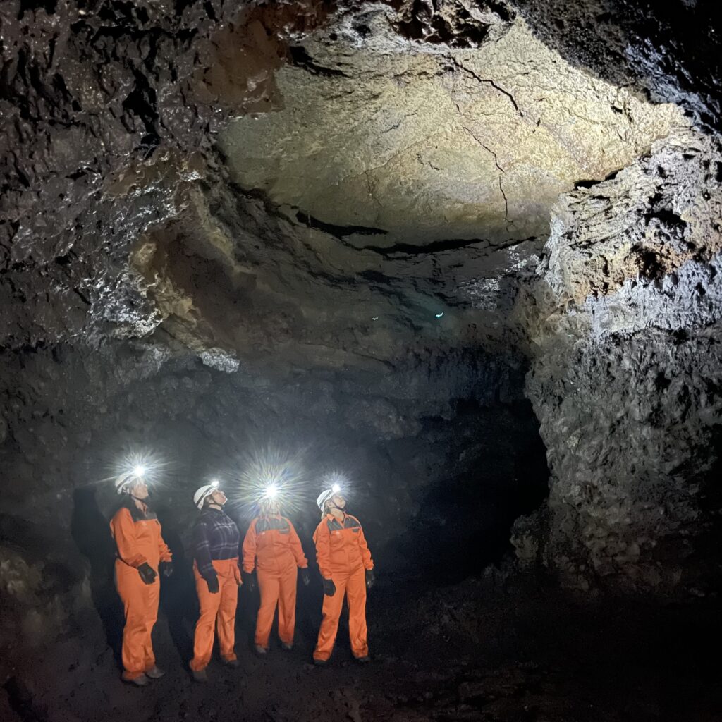 A group of four cavers wearing orange jumpsuits and helmets with headlamps explore a large lava cave, illuminating the rugged volcanic ceiling.