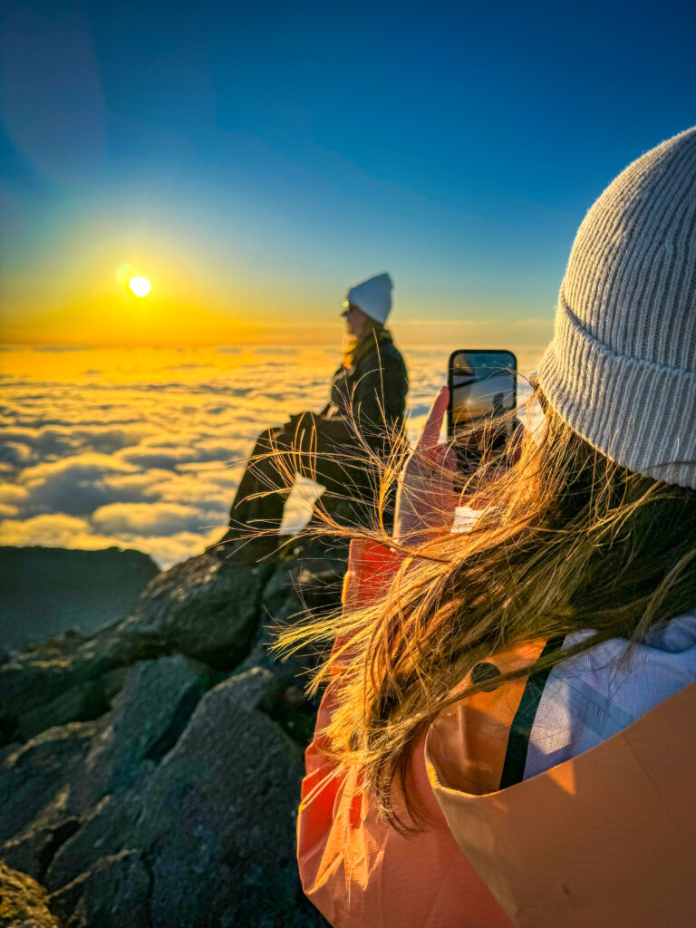 A hiker captures the golden sunset from the summit of Mount Pico, sitting above a sea of clouds.