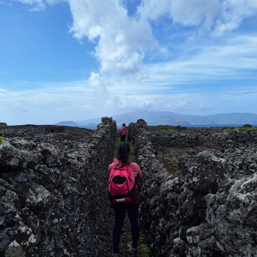 Two hikers walking along a narrow lava stone path on the west coast of Pico Island, Azores, with mountains and ocean in the background.