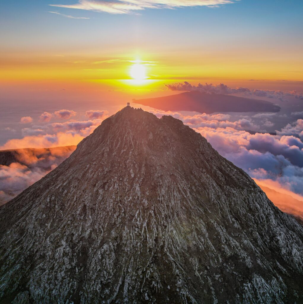 A breathtaking sunset view from the summit of Mount Pico, with two hikers standing at the peak above a sea of clouds.