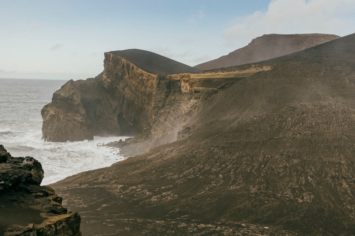 Rocky volcanic landscape with steep cliffs and ocean waves below under a partly cloudy sky.