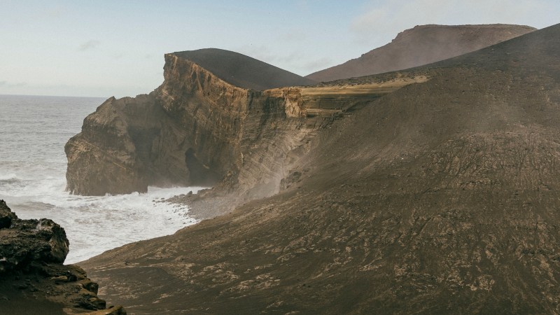Rocky volcanic landscape with steep cliffs and ocean waves below under a partly cloudy sky.