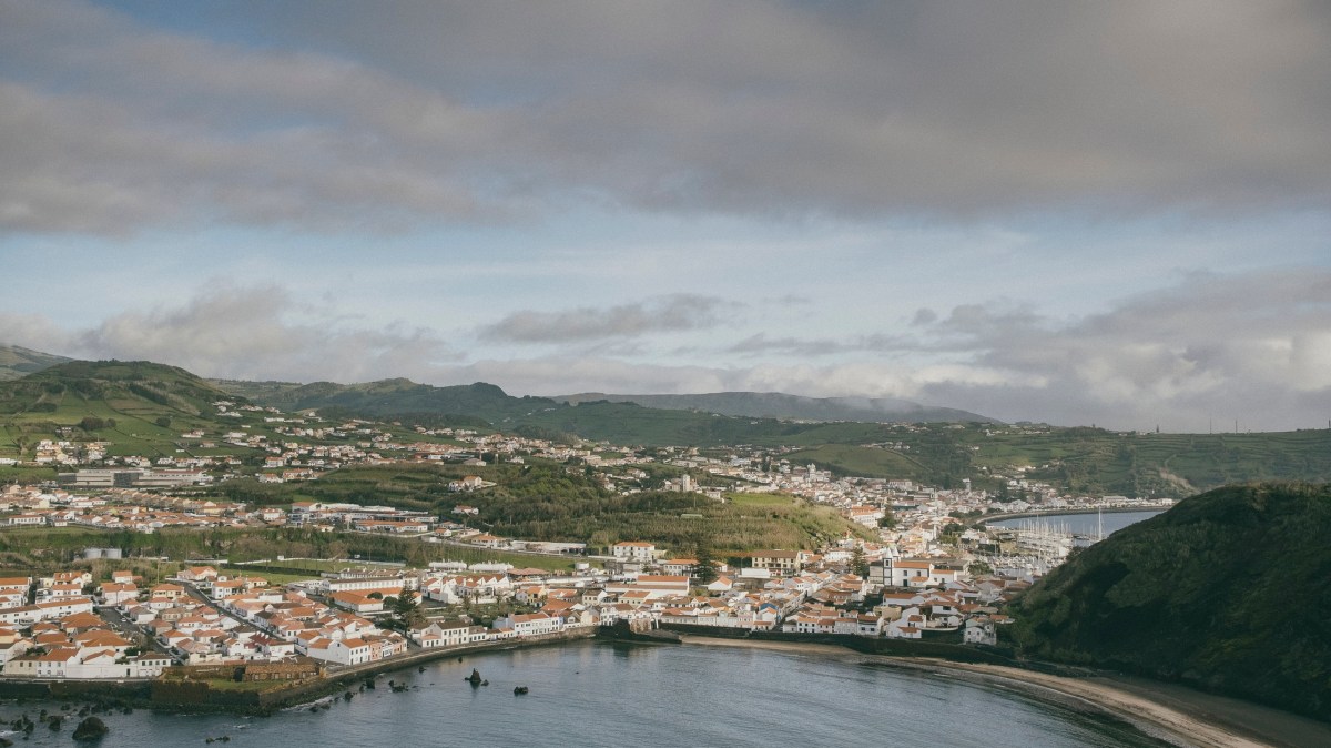 Coastal town with white houses and red roofs, surrounded by hills under a cloudy sky.