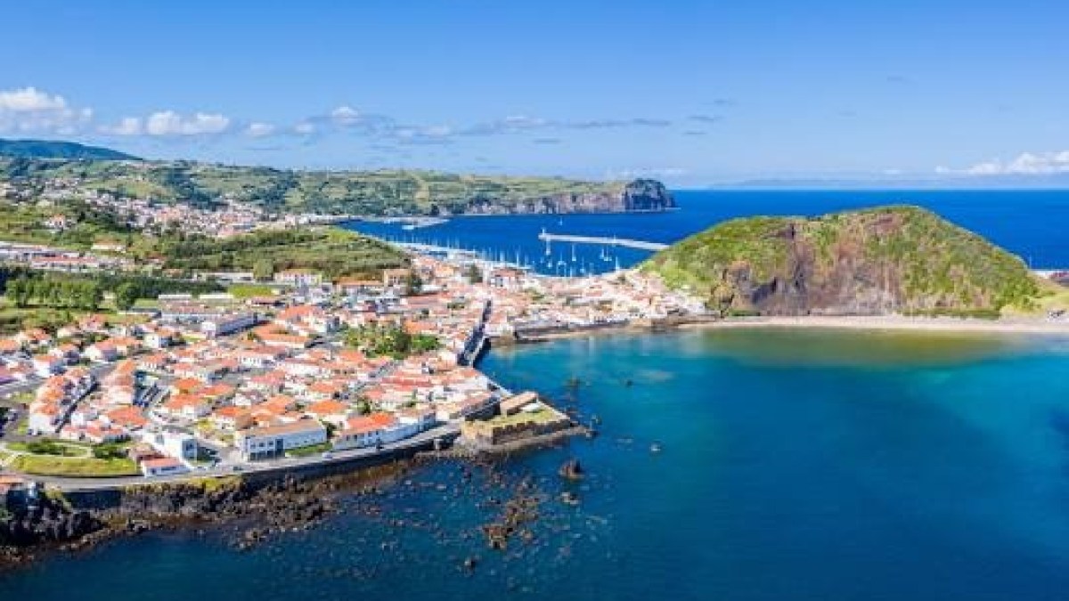 Aerial view of a coastal town with red-roofed buildings, green hills, and blue ocean under a clear sky.
