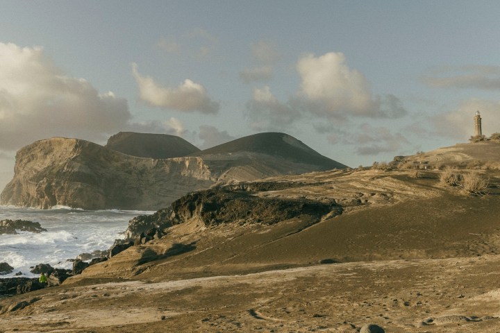 Rocky coastal landscape with hills, a lighthouse, and waves under a partly cloudy sky.