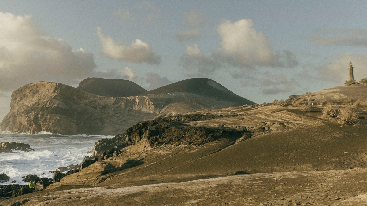 Rocky coastal landscape with hills, a lighthouse, and waves under a partly cloudy sky.