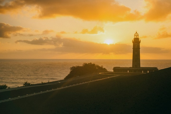 Sunset over ocean with lighthouse silhouette and road in foreground.
