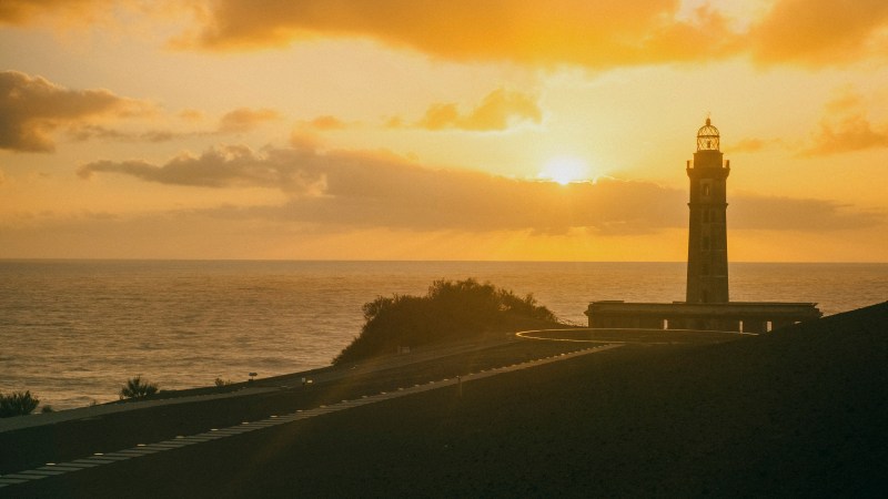 Sunset over ocean with lighthouse silhouette and road in foreground.