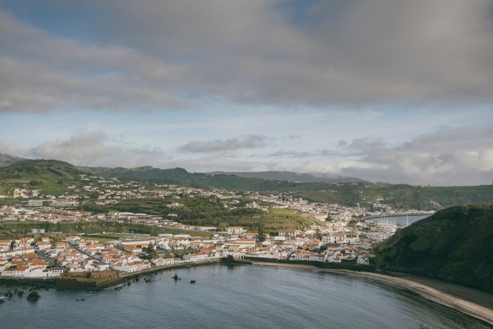 Coastal town with red-roofed buildings, green hills, and harbor under cloudy sky.