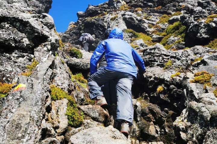 Person in blue jacket climbing rocky terrain under clear blue sky.