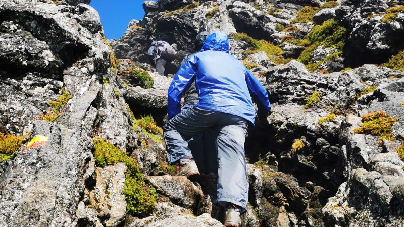 Person in blue jacket climbing rocky terrain under clear blue sky.
