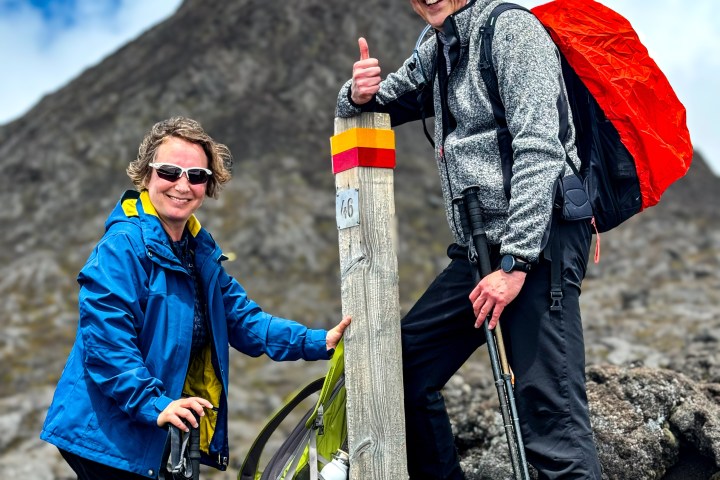 Two hikers smiling by a mountain marker with poles and backpacks in rugged terrain.