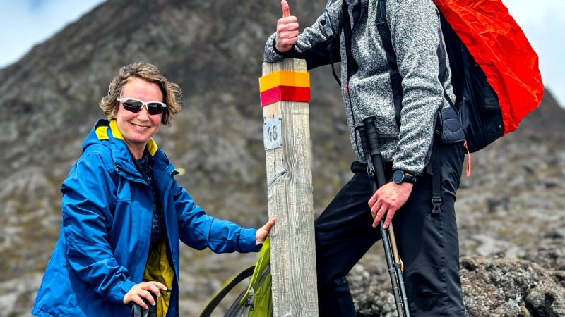 Two hikers smiling by a mountain marker with poles and backpacks in rugged terrain.