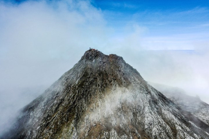 Peak of a rocky mountain shrouded in mist, with people standing at the summit under a clear blue sky.