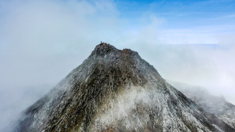 Peak of a rocky mountain shrouded in mist, with people standing at the summit under a clear blue sky.