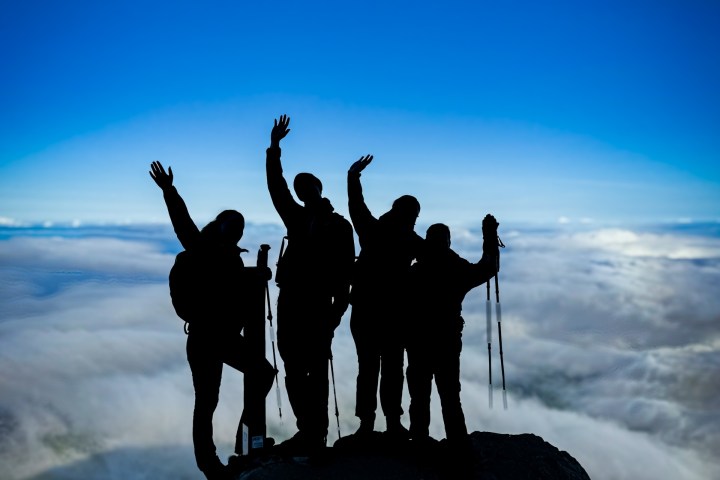 Silhouette of four hikers on a mountain peak against a cloud-filled sky.