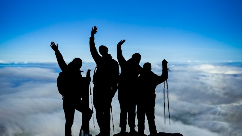 Silhouette of four hikers on a mountain peak against a cloud-filled sky.