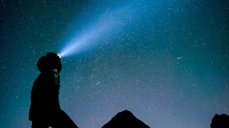 Silhouette of person with headlamp against starry night sky, standing on hill.