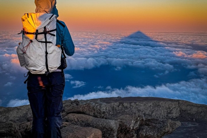Hiker with backpack faces sunset, clouds below, and mountain shadow on horizon.