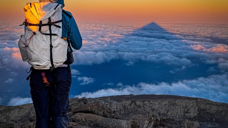 Hiker with backpack faces sunset, clouds below, and mountain shadow on horizon.