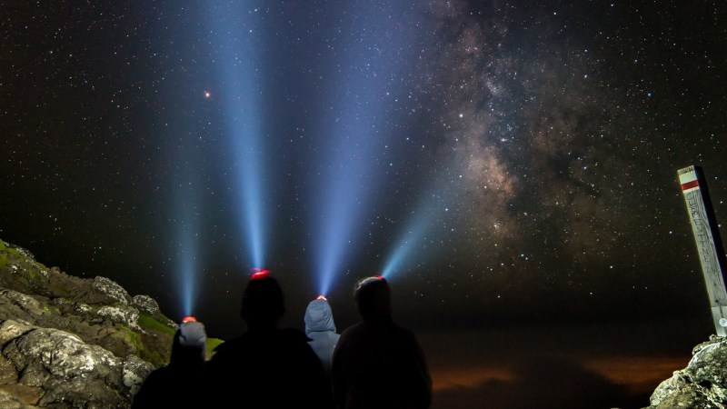 Three people with headlamps under a starry sky, beams pointing upwards towards the Milky Way.
