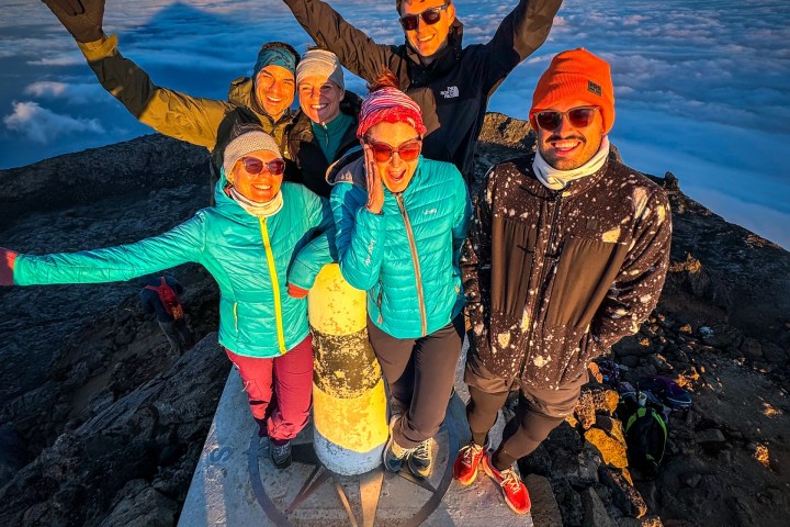 Group of six people celebrating on a mountain summit with a marker, above the clouds at sunrise.