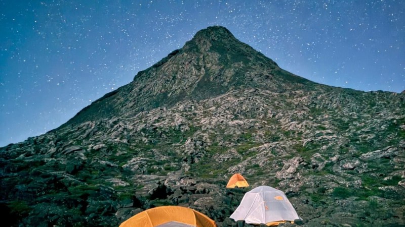Night scene with tents on rocky terrain beneath a starry sky and mountain.