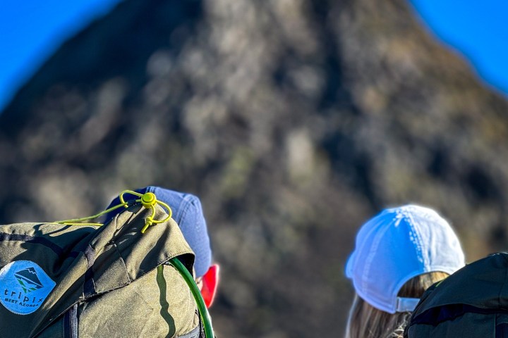 Two hikers with backpacks facing a large rocky hill under a clear blue sky.
