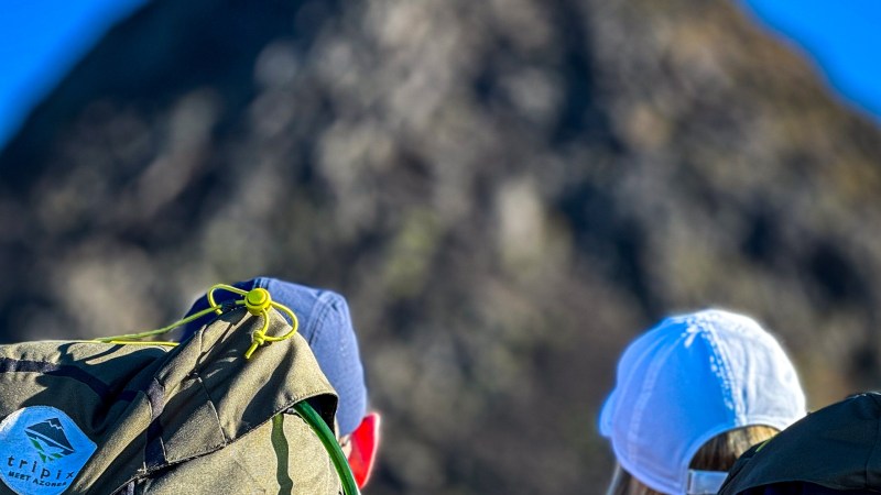 Two hikers with backpacks facing a large rocky hill under a clear blue sky.