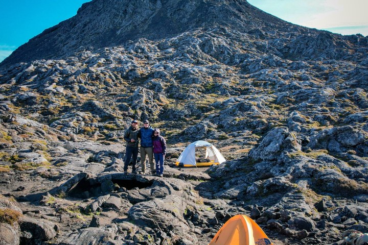 Three people and two tents on rocky terrain with a mountain in the background.