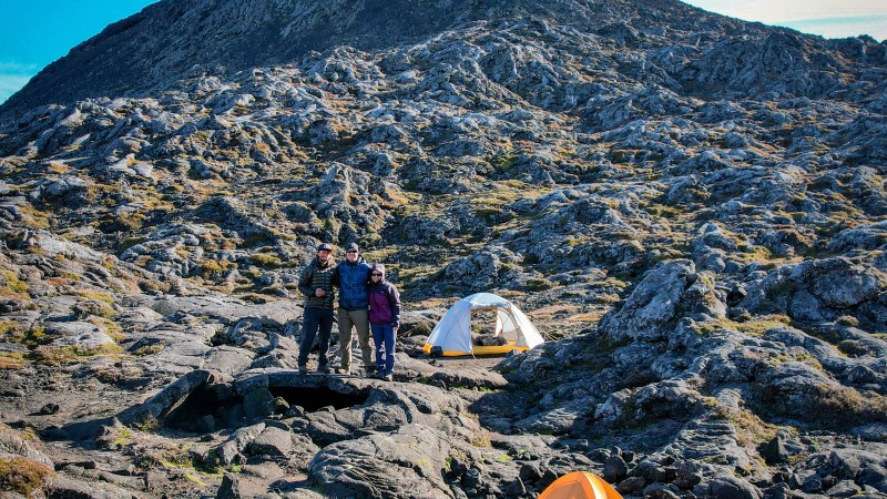 Three people and two tents on rocky terrain with a mountain in the background.