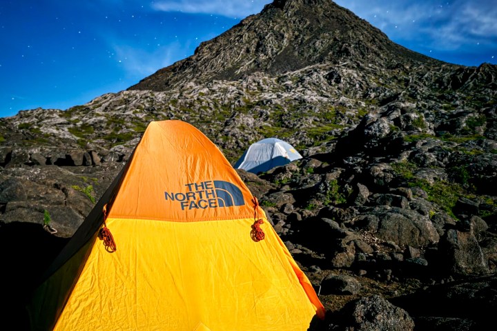 Orange tent on rocky ground under a starry sky with a mountain in the background.