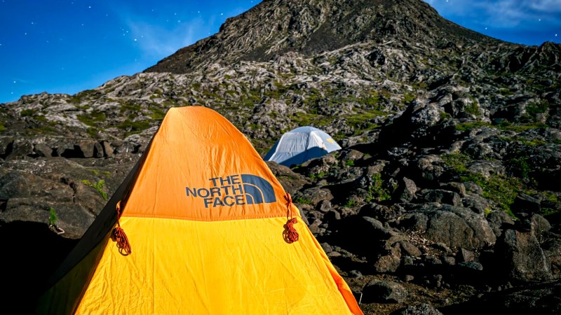Orange tent on rocky ground under a starry sky with a mountain in the background.
