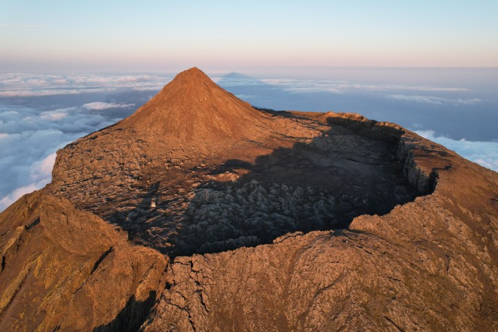 Aerial view of a volcanic mountain peak with a crater, surrounded by clouds.