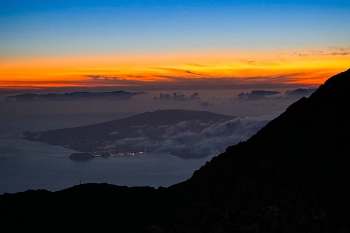 Mountain silhouette at sunset with orange sky and distant island view.