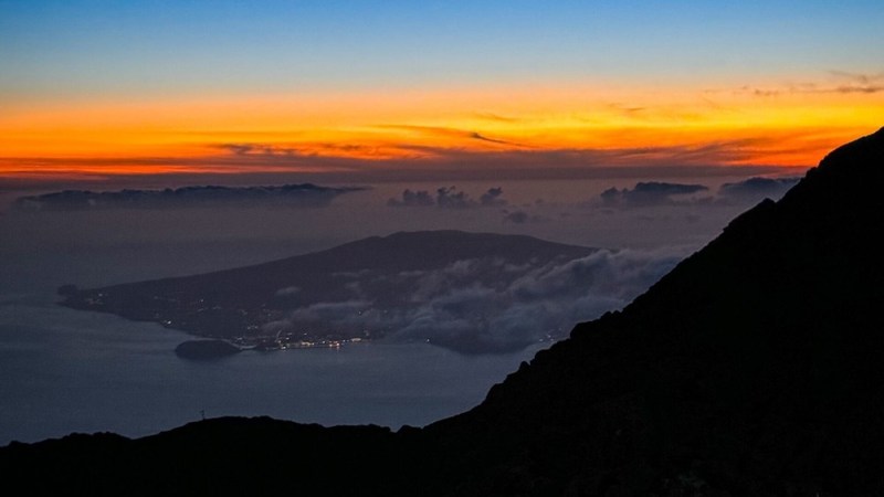 Mountain silhouette at sunset with orange sky and distant island view.