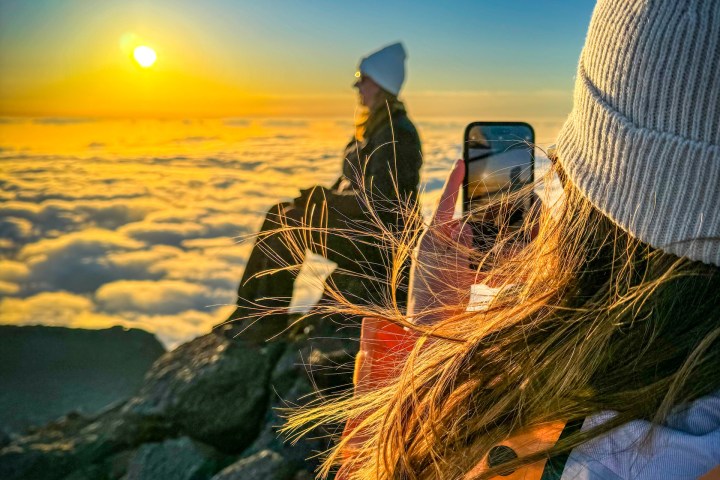 Two people on mountain summit at sunrise, one taking a photo, surrounded by clouds.