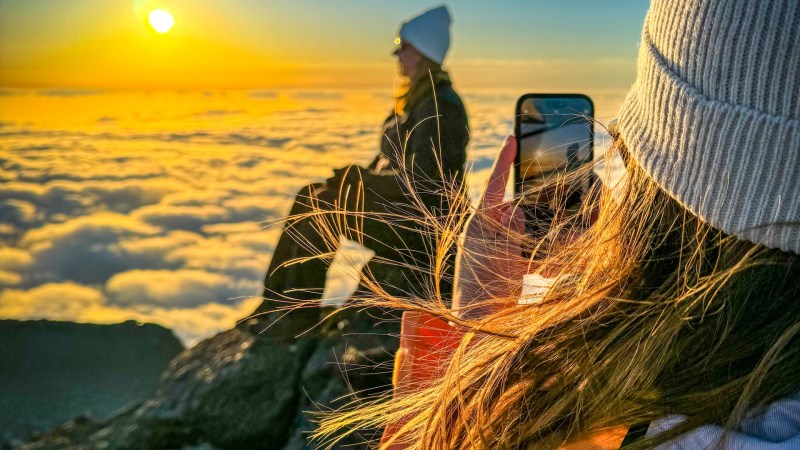 Two people on mountain summit at sunrise, one taking a photo, surrounded by clouds.