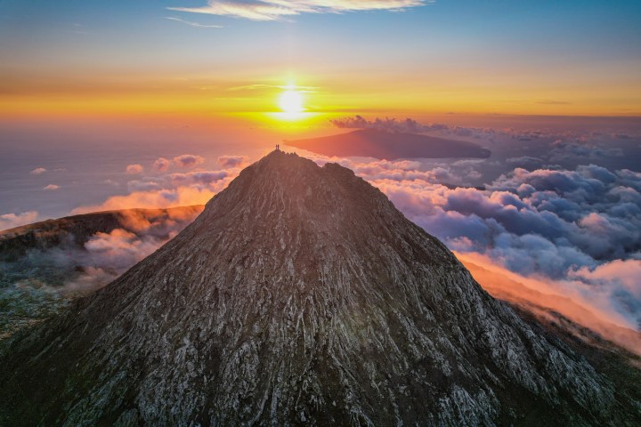 Mountain peak at sunrise with clouds and ocean in the distance, sky lit in warm colors.