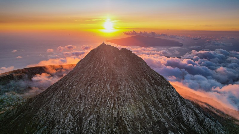 Mountain peak at sunrise with clouds and ocean in the distance, sky lit in warm colors.