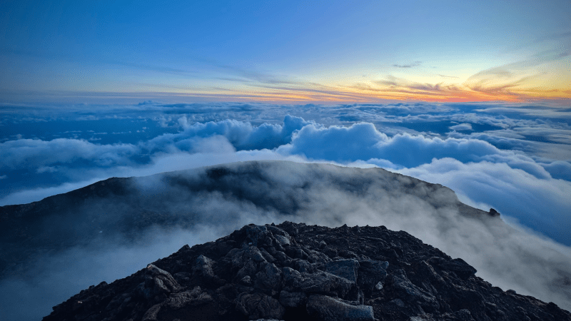 Rocky mountain peak above clouds at sunset, blue sky and horizon.